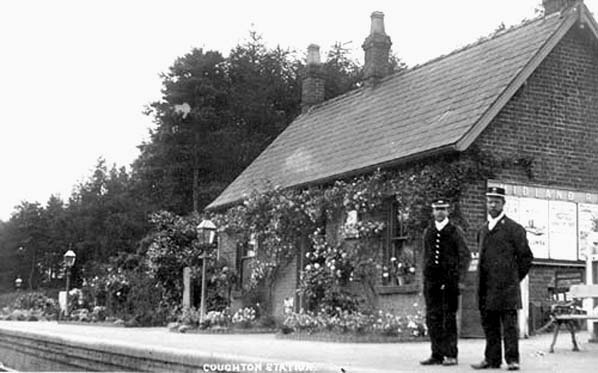 View of the simple station building bedecked with flowers with the Station Master and a porter posed on the platform