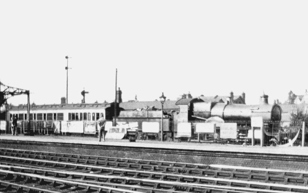 An unknown GWR 4-4-0 County class locomotive carrying express code lamps passes behind the station with a single coach