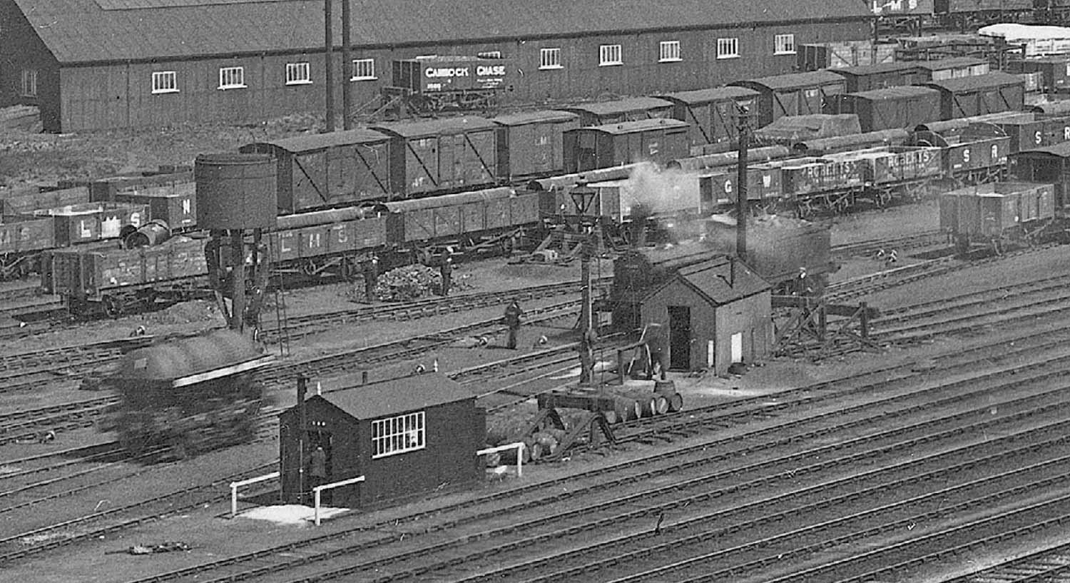 Close up showing an oil wagon being fly shunted passing the 'parachute' water column with the locomotive paused by the offices and mess huts of the yard staff